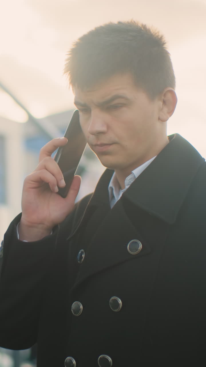 검은 코트를 입은 남자가  에 드폰을 들고, 불만을 표시하면서 전화 중에 세 번 니다. 배경에는 현대적인 유리 건물, 부드러운 빛 및 도시 풍경이 포함되어 있습니다.