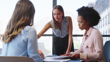 Female-Multi-Cultural-Business-Team-Meet-Around-Boardroom-Table-With-Laptops-Discussing-Documents