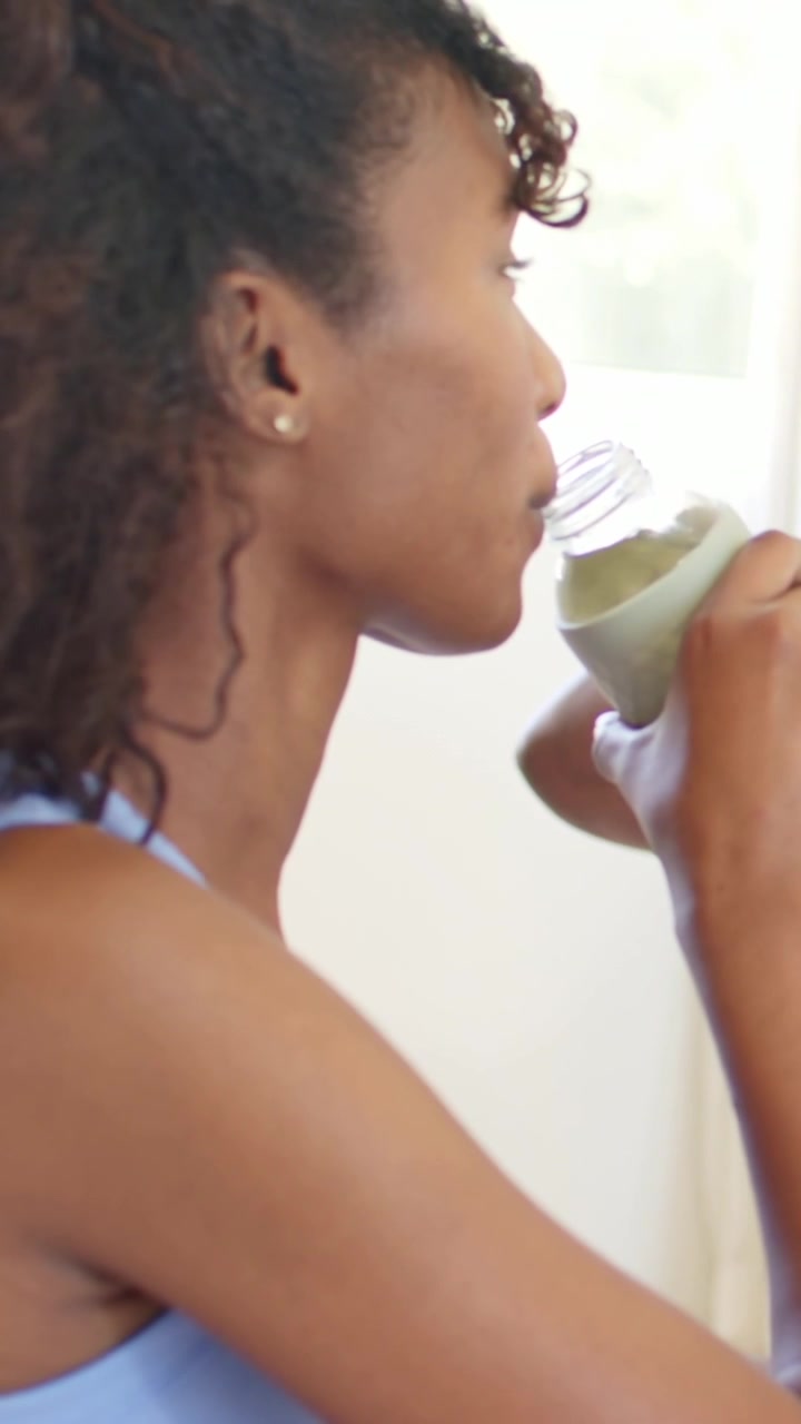 Lodret video: Kamera følger kvinde i lyseblå top, løfter grøn flaske og nipper ved vinduet