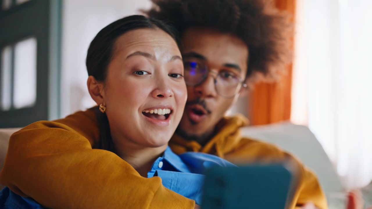 Couple taking selfie on sofa at home. Closeup of smartphone in mixed race hands.