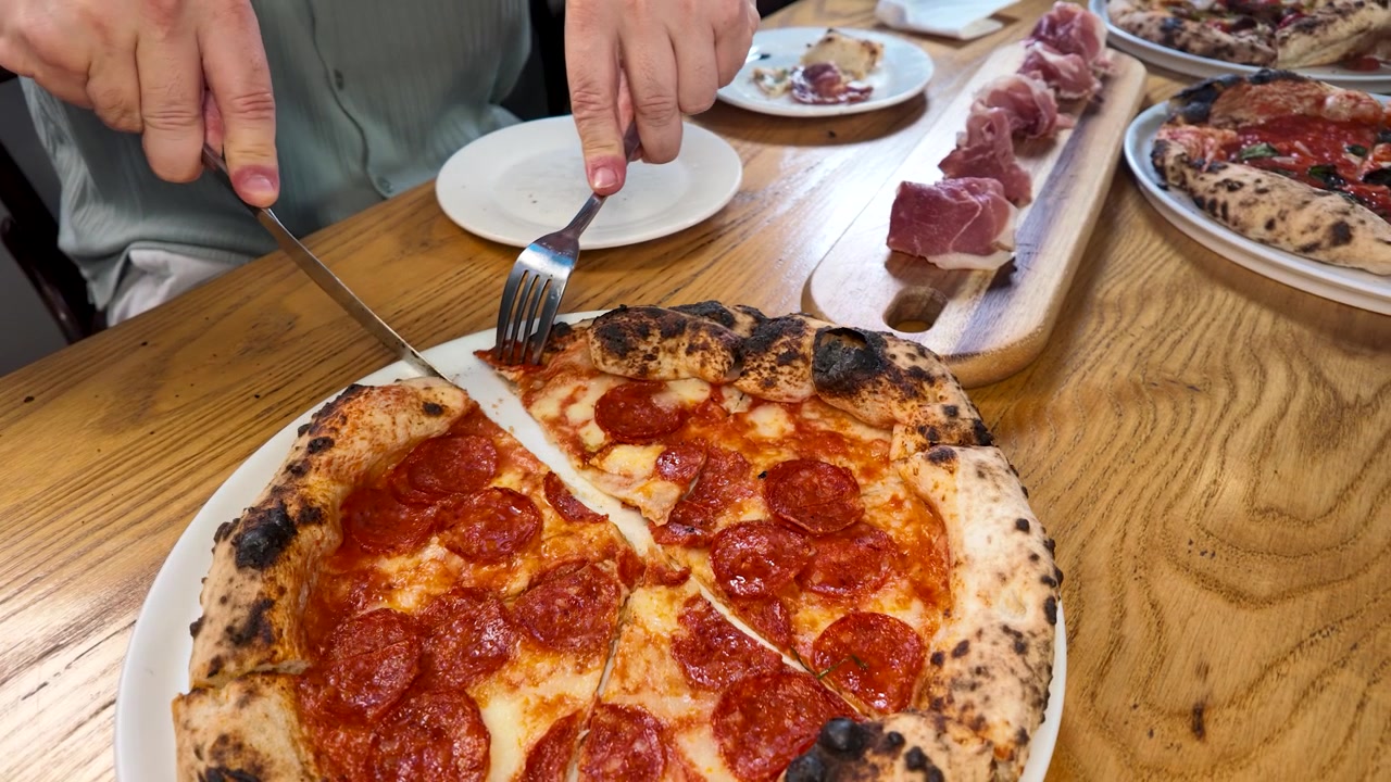 Primo piano di mani che tagliano una pizza al salamino piccante cotta nel forno a legna su un tavolo di legno con salumi.