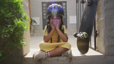 happy african american girl wearing helmet sitting on stairs with skateboard and looking at camera