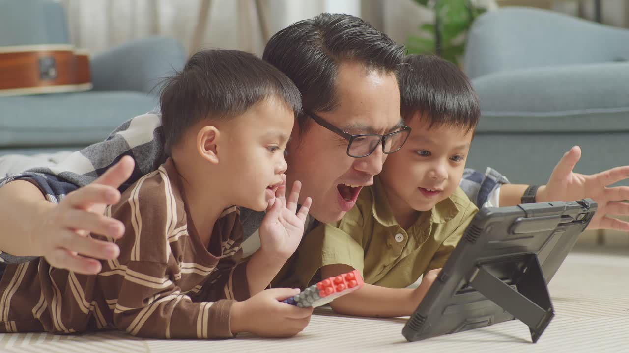 플라스틱 장난감 벽돌을 가지고 바닥에 누워있는 아시아 아버지와 아들의 근접 사진, 집에서 함께 태블릿에서 영화를보고, 아버지가 비명을 지르며 골을 축하합니다.