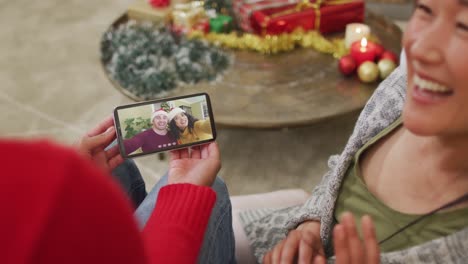 pareja asiática sonriente usando un teléfono inteligente para una videollamada de navidad con pareja con sombreros de papá noel en la pantalla