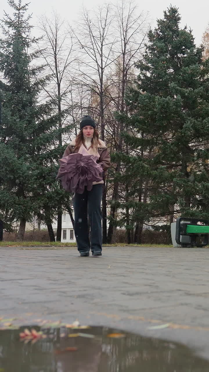 Female walking peacefully on cold overcast day through paved park holding closed umbrella, dressed in black knit cap, brown shearling jacket, and black trousers, gentle snowfall