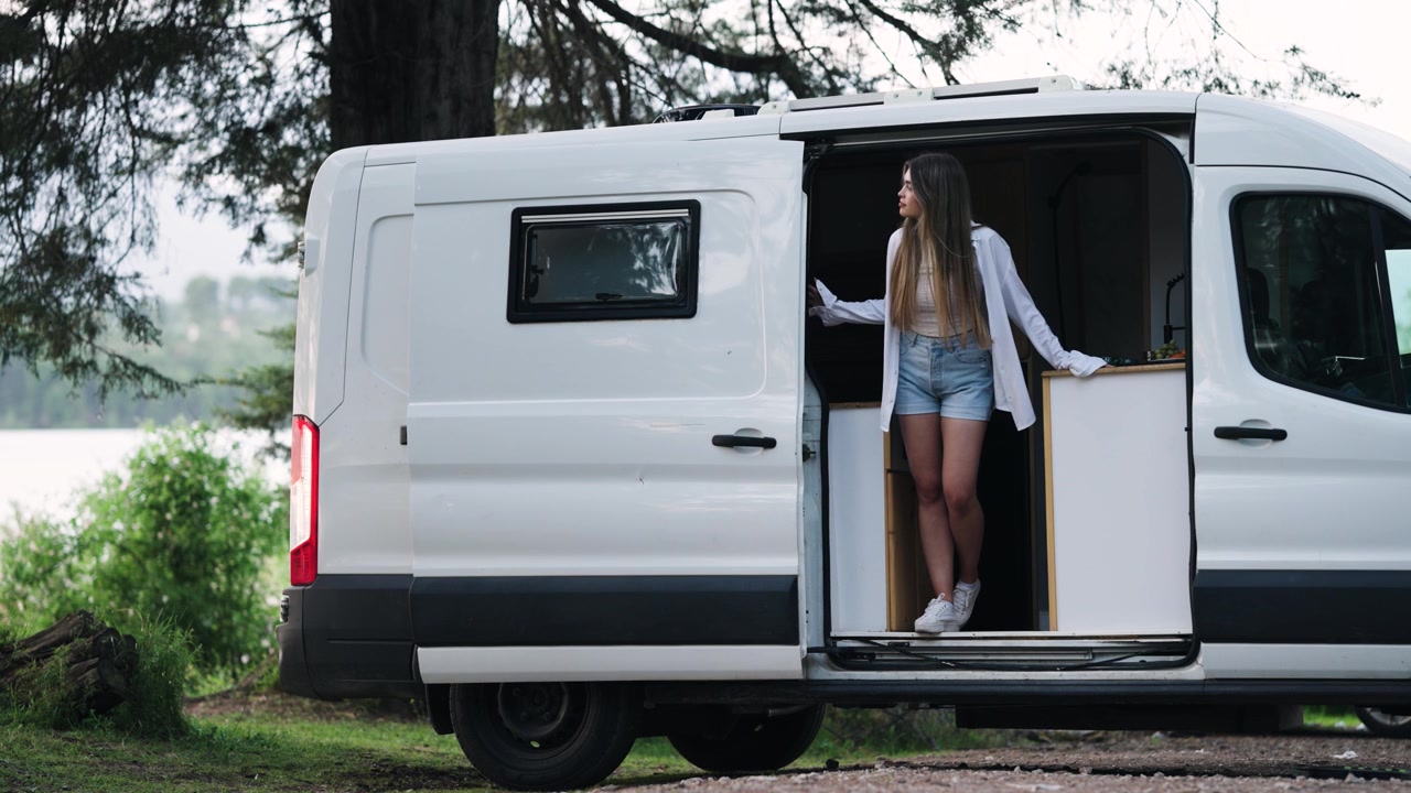 Giovane donna in piedi sulla porta aperta di un camper che guarda il lago