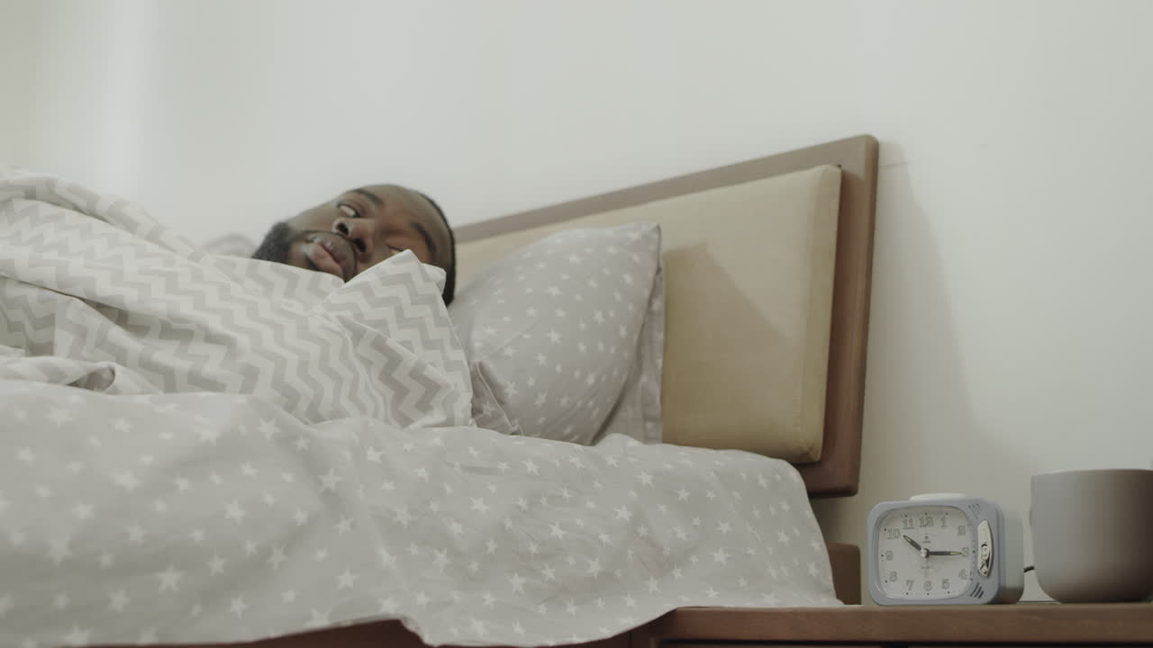 hombre negro apagando la alarma por la mañana. joven adulto despertando en la cama lentamente.