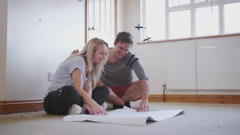 couple sitting on floor looking at floor plans in empty room of new home