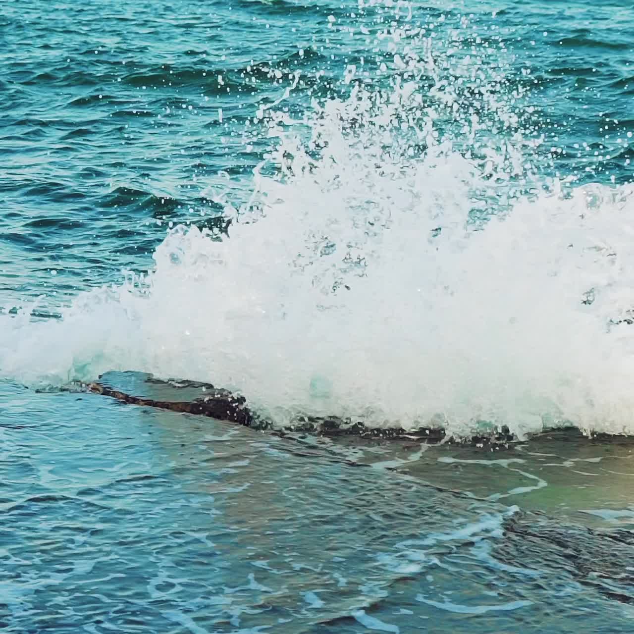 Turquoise waves is colliding with stone rocks near the shore and turning into thousands of drops of water. Slow motion. View from above