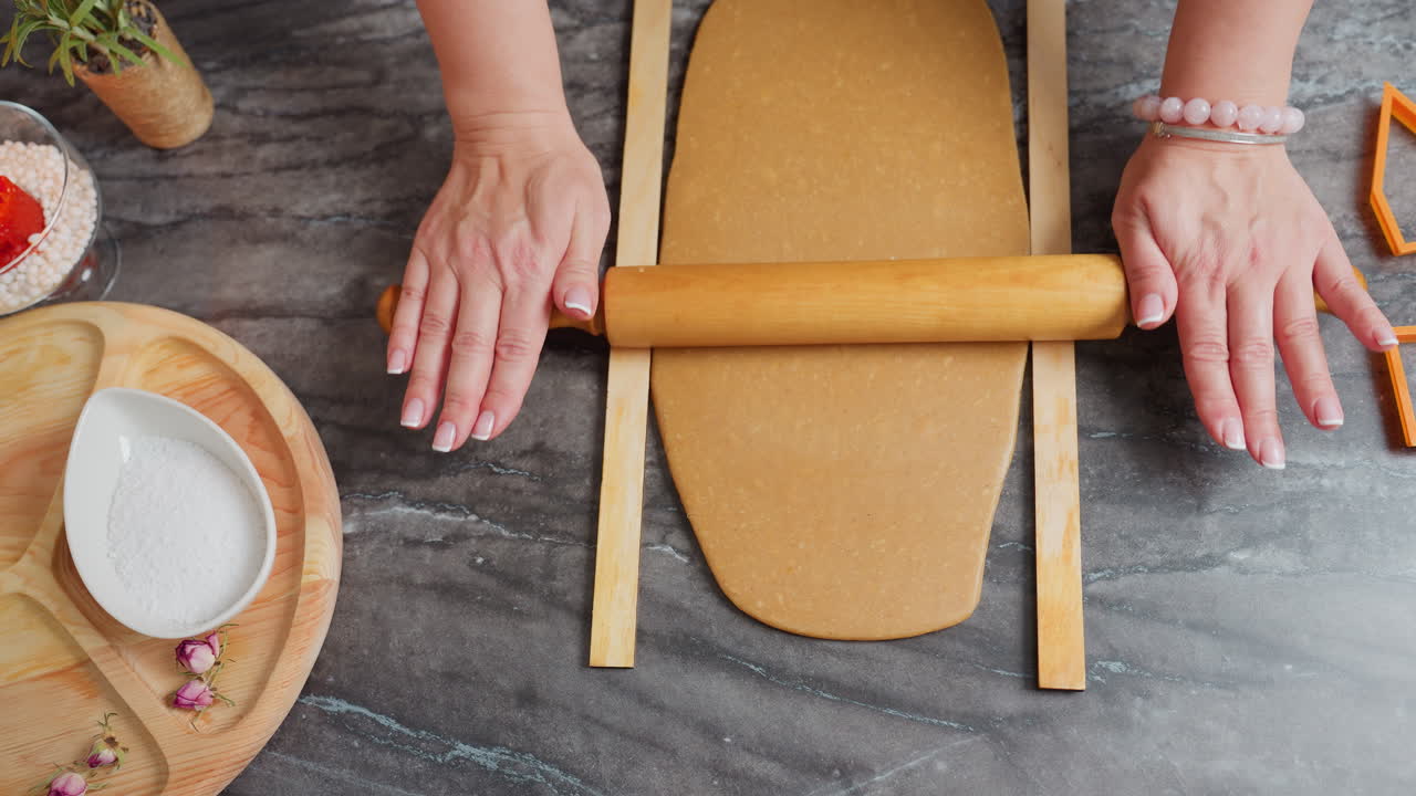 vista de arriba hacia abajo de un panadero rodando la masa con un rodillo de madera, manos presionando cuidadosamente, pulsera de cuentas rosas en la muñeca, rodeado de herramientas de horneado, bandeja de madera con sal, flores secas
