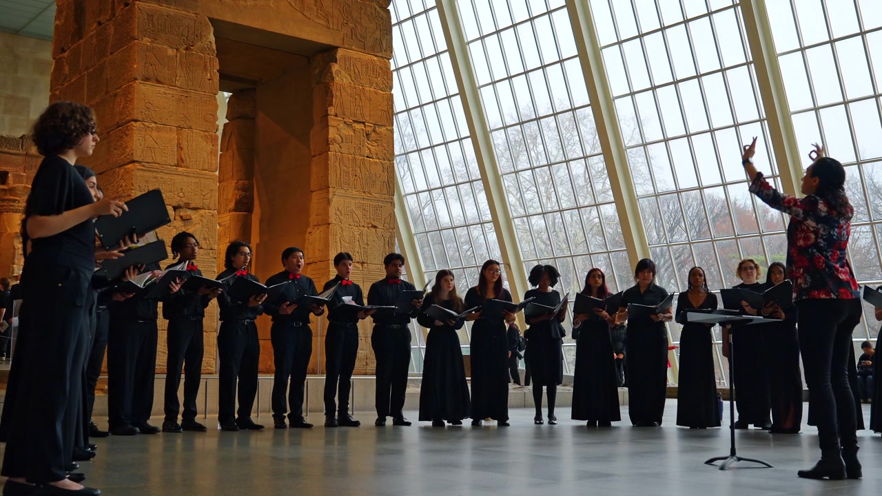 New York, USA, 18 marzo 2026: Ensemble vocale che canta in un sito di un antico tempio egizio in un museo. Coro multietnico in abiti neri che si esibisce con un direttore davanti a un tempio egizio in pietra storico.