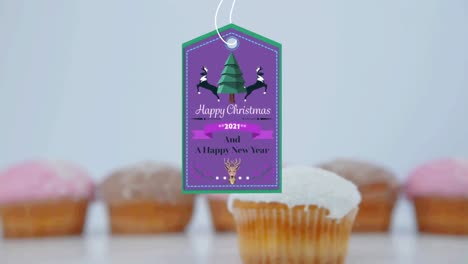 Animation-of-christmas-greetings-on-tag-over-cupcakes-on-white-background