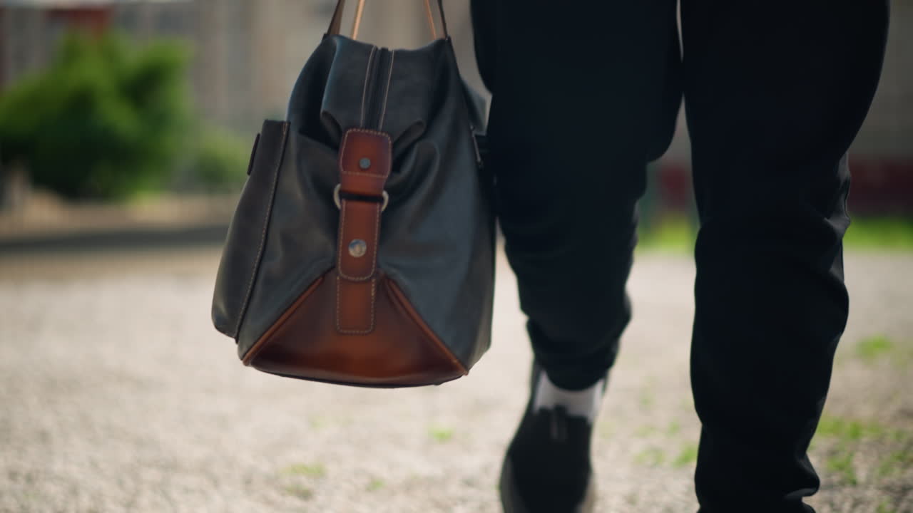 hombre paseando al aire libre, individuo con una bolsa caminando al aire libre, hombre cargando una bolsa de cuero en un tranquilo parque, figura masculina informal paseando por un sendero de grava en un parque al amanecer con una bolsa balanceándose