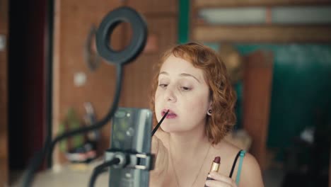 beauty blogger redhead woman applies lipstick on camera, influencer filming herself doing make-up with smartphone