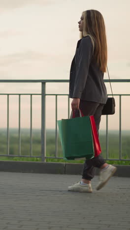 vista lateral de una dama con ropa gris caminando a lo largo de un carril de hierro con una bolsa de compras y un bolso de mano negro, fondo borroso con un banco de piedra y vegetación distante bajo una iluminación suave y tranquila al amanecer