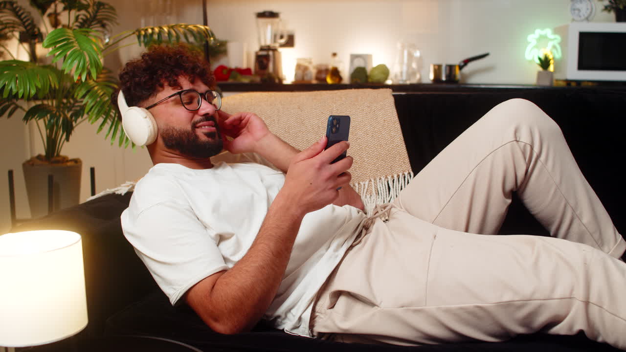 Arabian man with wireless headphones listens to music chats on smartphone at night indoors relaxed