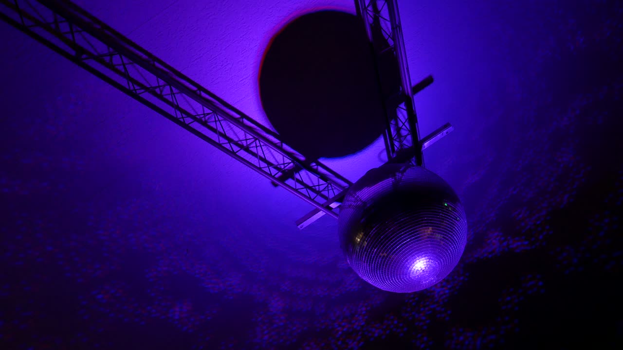 Footage of a spinning mirrored sphere disco ball with reflections by colorful lights on the roof.