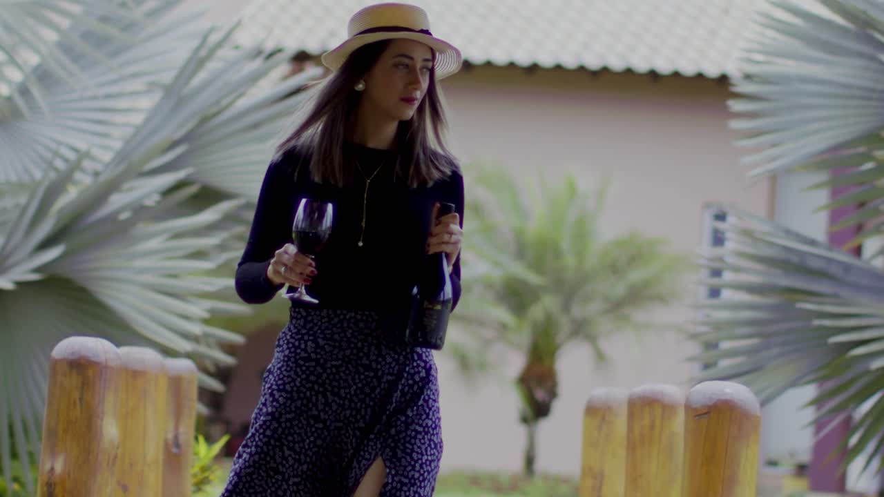 señora que lleva un sombrero de paja camina en un jardín tropical con un vaso y una botella de vino tinto en sus manos - cierra a cámara lenta