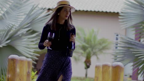 lady-wearing-a-straw-hat-walks-in-a-tropical-garden-with-a-glass-and-a-bottle-of-red-wine-in-her-hands---close-up-slow-motion