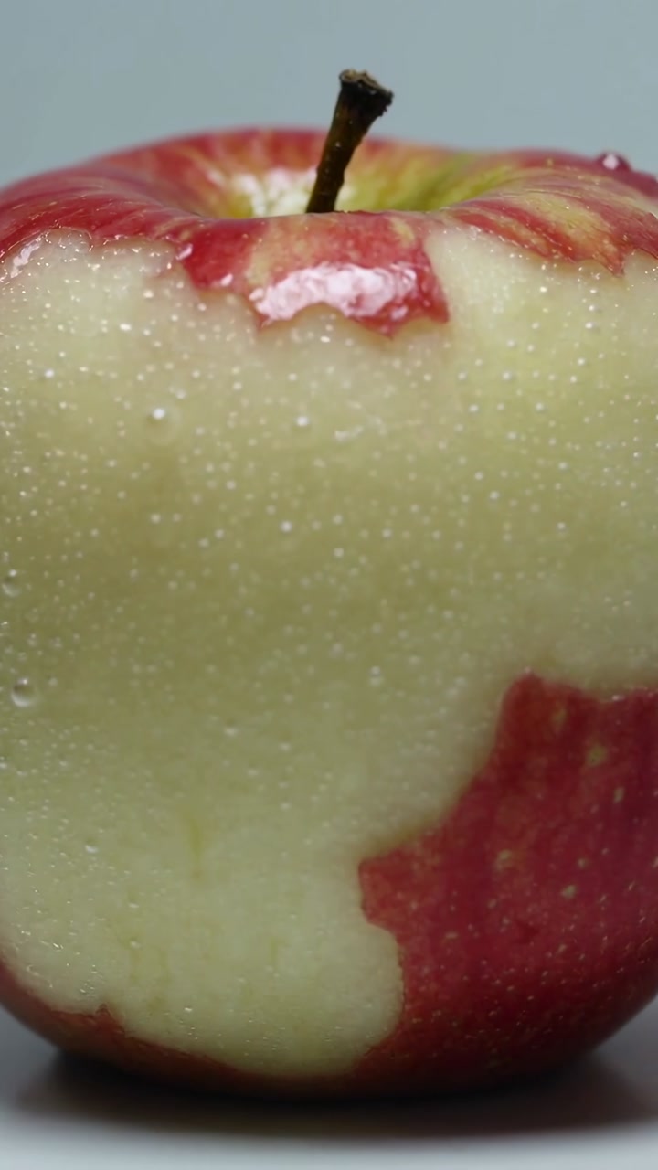 Vertical video: Advancing frames showing red apple stem losing peel on gray table, exposing flesh
