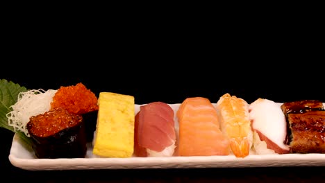 a rectangular plate with assorted sushi nigiri, sashimi, and tamago rotates smoothly against a black background under soft, even lighting