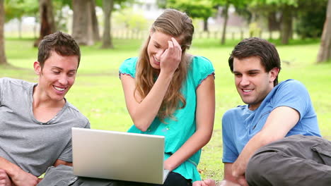 Drei-Freunde-Unterhalten-Sich,-Während-Sie-Auf-Einen-Laptop-Schauen,-Während-Sie-In-Einem-Park-Sitzen