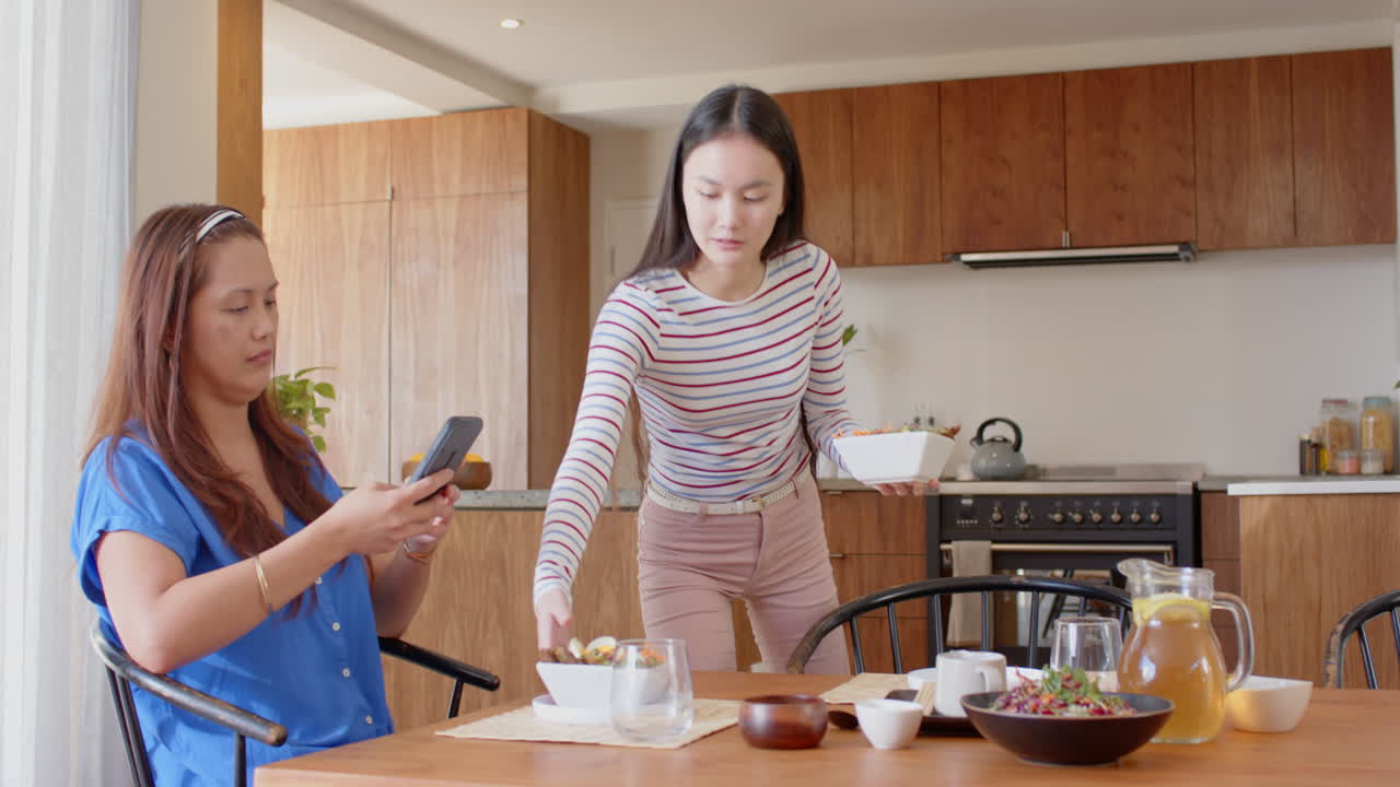 Setting table for meal, young woman while older woman using smartphone in kitchen