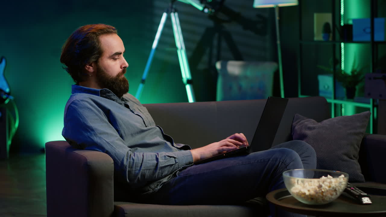 hombre trabajando en una computadora portátil en casa con un programa de televisión jugando en el fondo