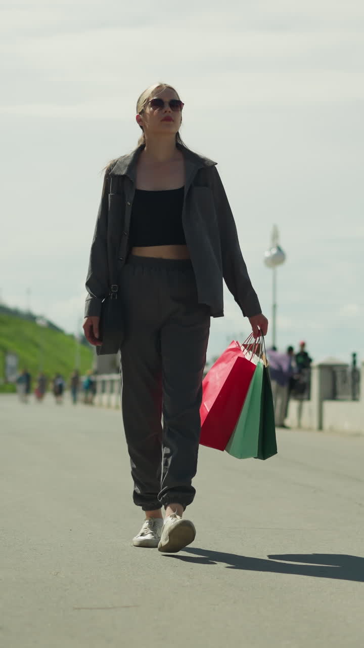 dama con sombrillas de sol sosteniendo bolsas de compras rojas, menta y verdes, mirando hacia abajo mientras camina y luego mirando hacia otro lado, su sombra se ve en el camino asfalto, el fondo es borroso con otras personas