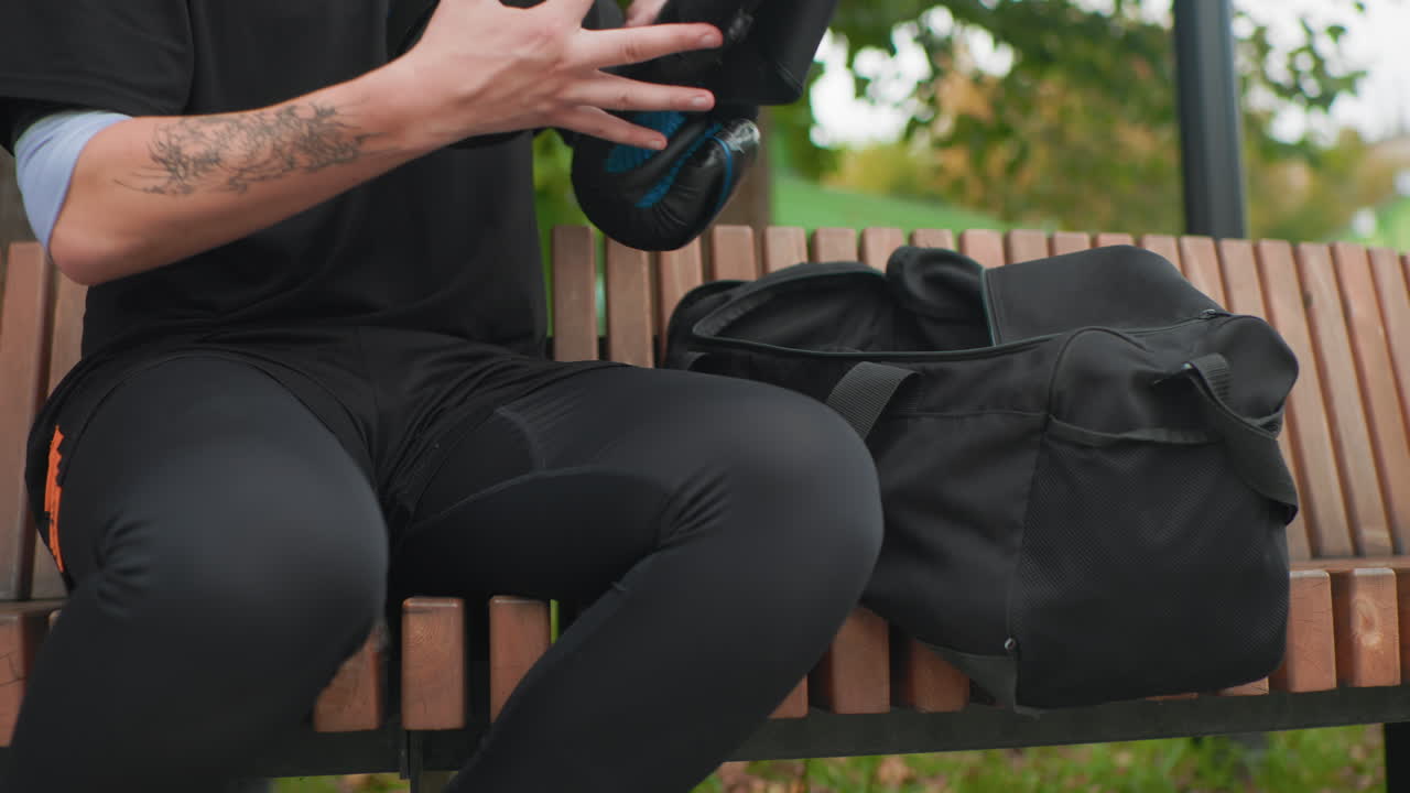 Ein weißer Mann sitzt auf einer Bank, greift in eine schwarze Tasche und entdeckt einen Fahrradhelm, tätowierter Unterarm sichtbar, Parkbäume im Hintergrund, lässige Pendlerpause mit konzentrierten Händen, die Schutzausrüstung herausziehen