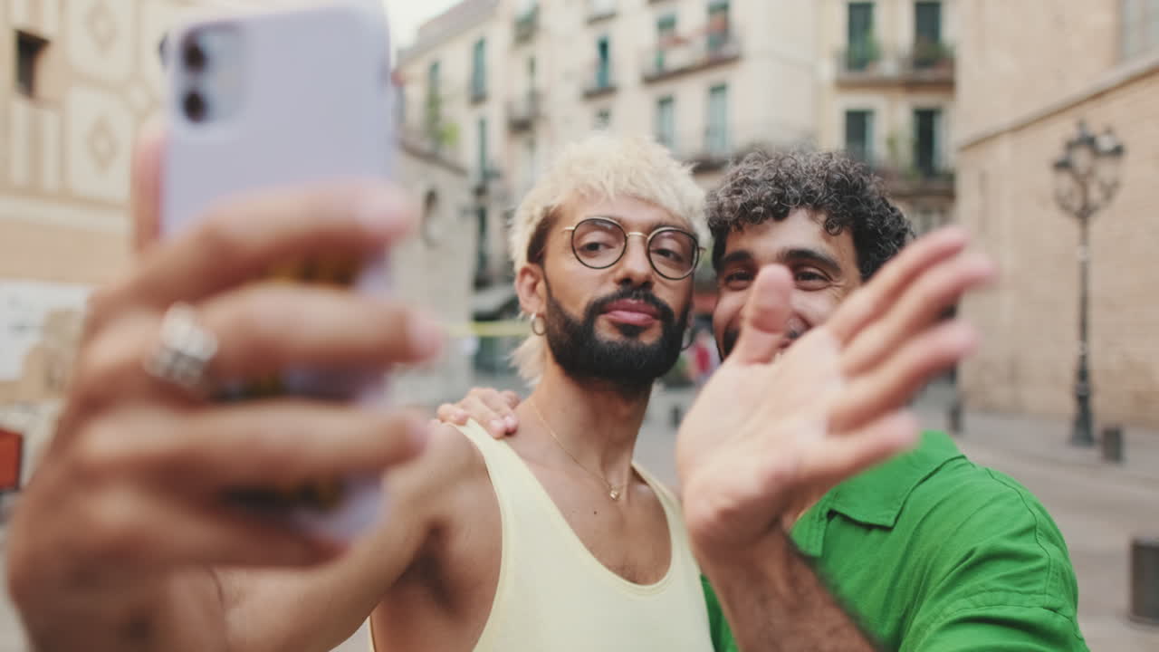 Giovane coppia gay felice che si scatta un selfie e saluta in una strada di città