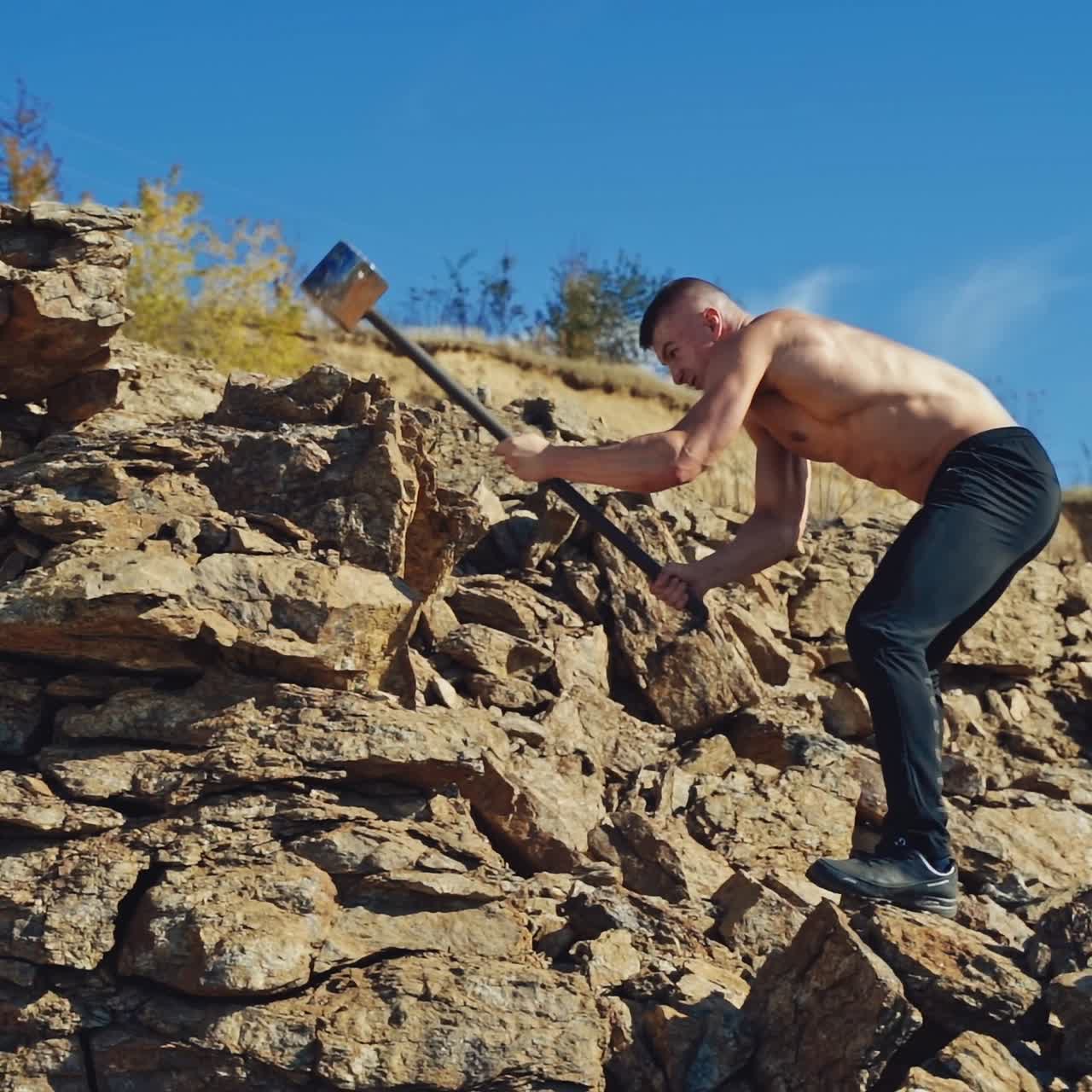 Side view of a strong man breaking stones with a hammer. Shirtless athletic man exercising with a sledgehammer on the rocky background. Slow motion.