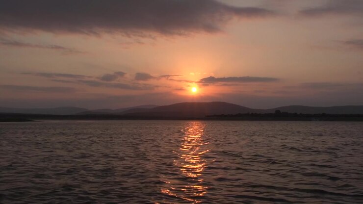 Spectacular wide shot of a glowing golden sunset over a large rippling lake, with sun rays bursting through clouds behind a beautiful silhouette of rolling mountain ridges in South Africa.