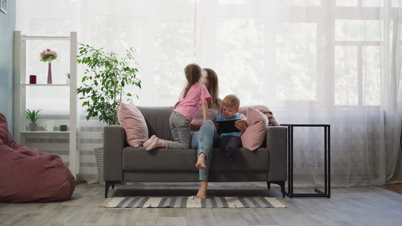 la niña besa a la madre mientras el niño pequeño ve dibujos animados en la tableta