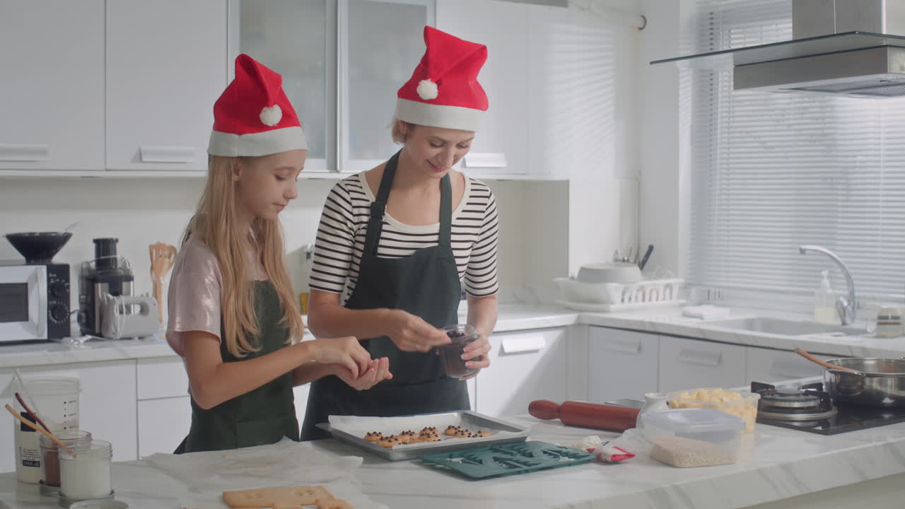 Girl and Mother in Christmas Hats Adding Chocolate Chips to Cookies