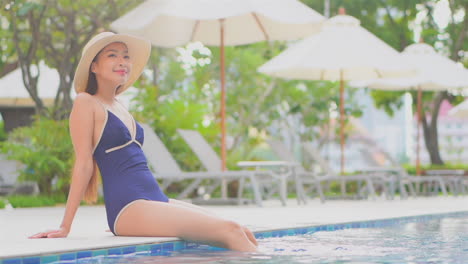 Asian-girl-sitting-on-pool-edge-with-swimsuit-and-wide-brimmed-hat