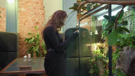 woman stands by glass terrarium smiling softly while taking photo of lush greenery inside, near reserved table with ceramic d cor, in cozy indoor setting with exposed brick and potted plants