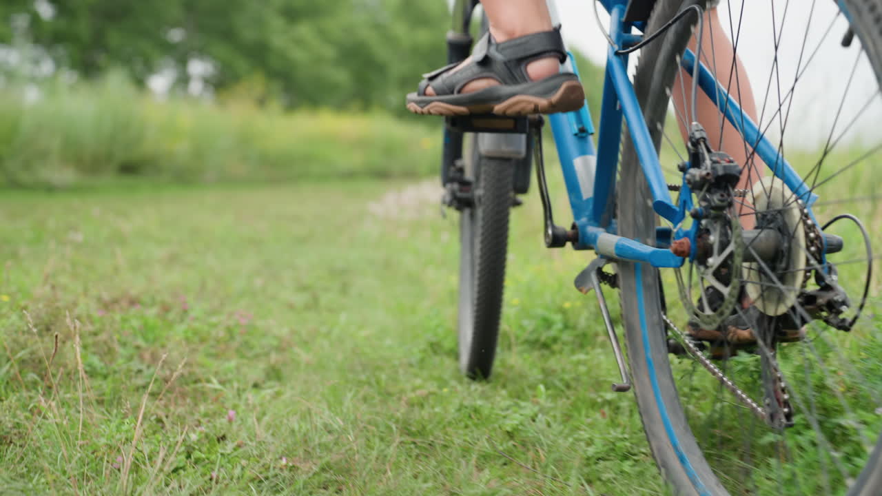 파란색 자전거를 탄 흰색 머리 소년이 들판과 수평선을 바라보고 있어요. 편안한 자세로 샌들을 페달에 올린 채 안정적인 바퀴를 클로즈업하고 넓은 초원을 배경으로 부드러운 바람이 불어오네요. 라이딩 전 잠시 생각에 잠긴 듯한 모습에 은은한 흐린 날씨의 빛이 감싸고 있어요.