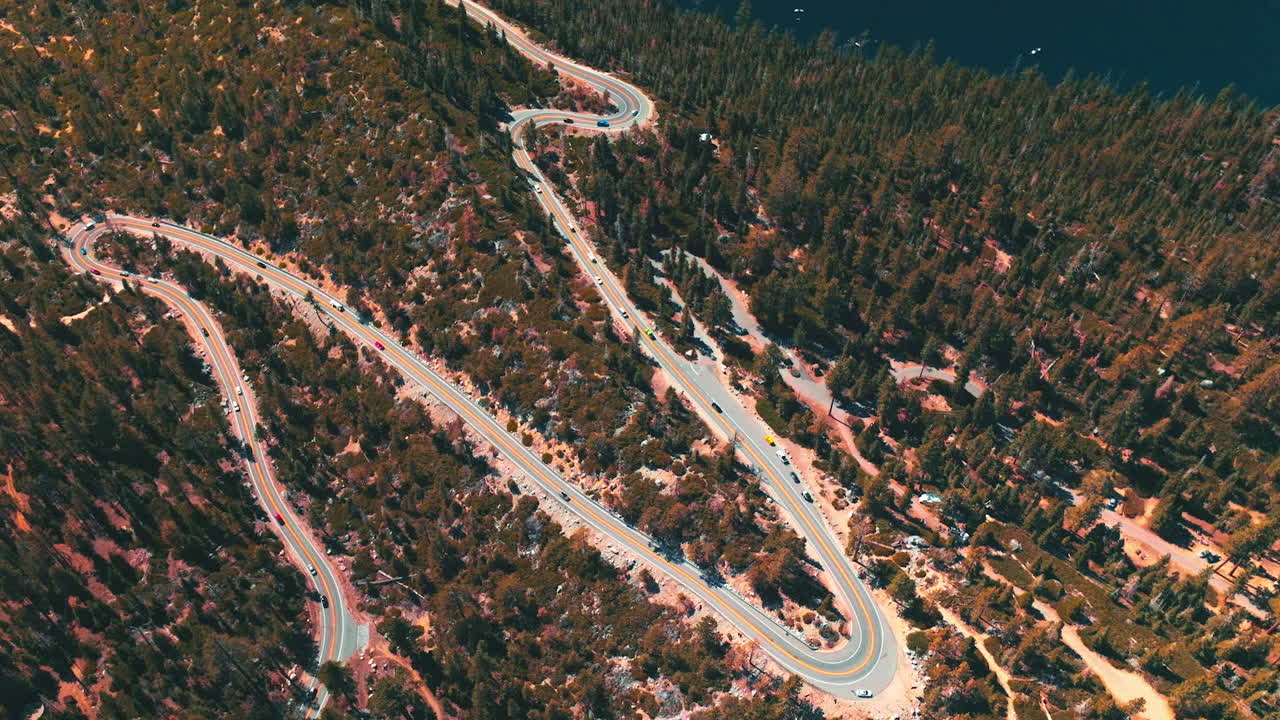 Twisted road through the wooded rocky landscape with many cars moving by. Deep blue lake at the foot of mountain from aerial perspective.