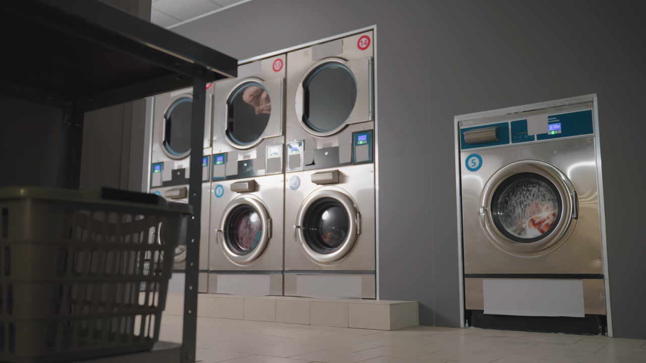 Zoom out on labelled steel washing machines in row spinning clothes, unit by corner, laundry baskets visible, steel doors, timers glowing, self service laundromat, wash cycle motion behind glass