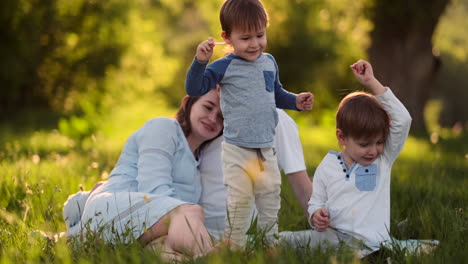 dad mom and two sons in the summer at sunset sitting in a meadow on the grass laughing and hugging each other.