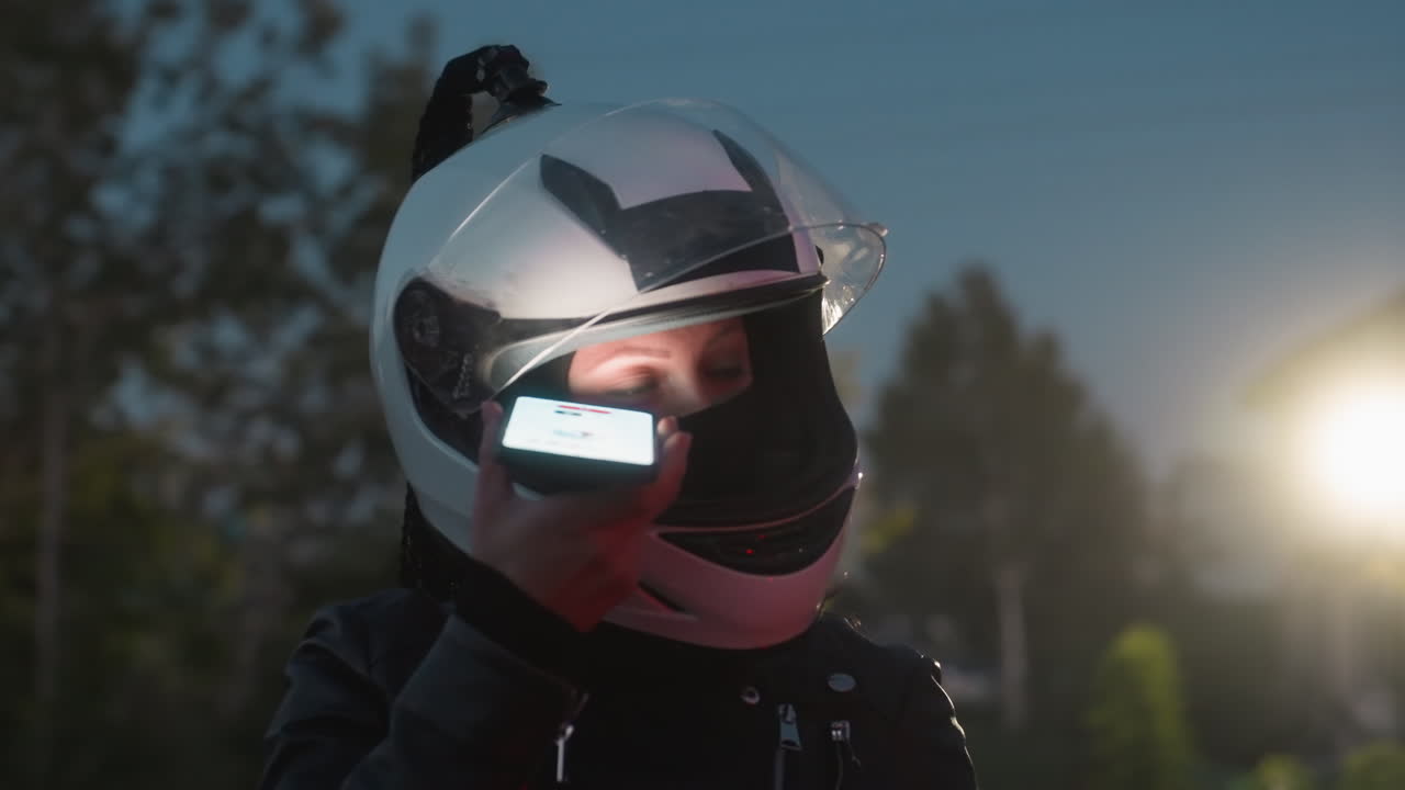 Vizörü kalkık bir kask takan kadın, akşam saatlerinde yol kenarında durmuş telefonla konuşurken, arkadan ışıkları yanan bir araba geçiyor; yeşillikler ve bulanık farlar görülüyor.