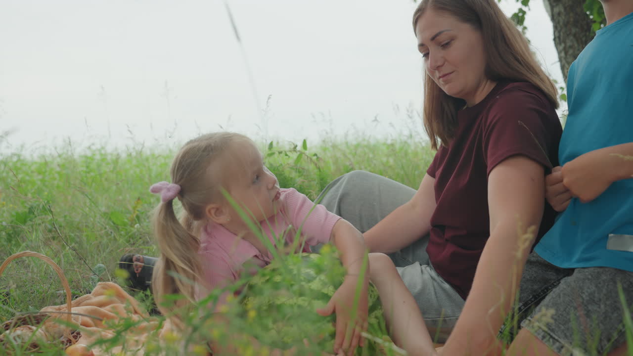Familia blanca recogiendo sandías en el prado, la madre enseña a su hija a sujetar la fruta mientras el hermano mayor inspecciona la corteza, manos ocupadas con la cesta y la manta, risas alegres, lección de cosecha, ayudante de granjero