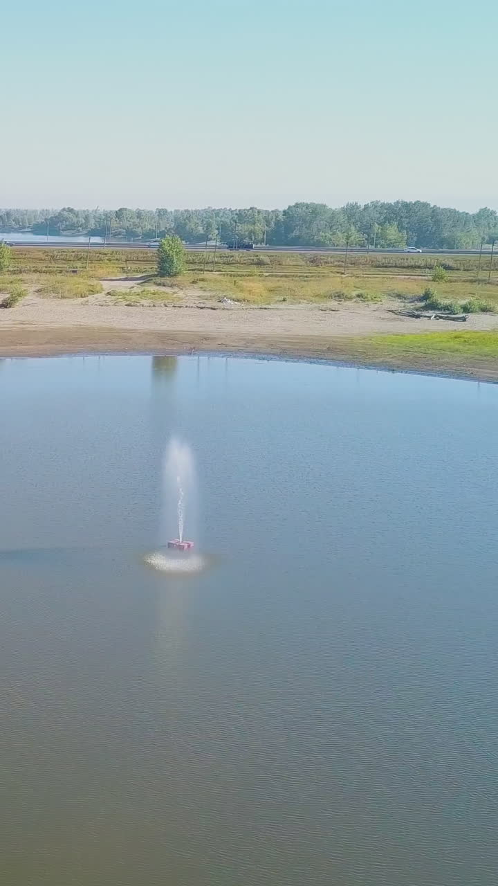 small jet fountain in wide calm lake middle among green meadows against clear blue sky on sunny day upper view