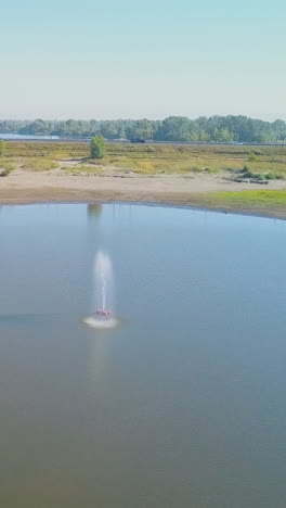 small jet fountain in wide calm lake middle among green meadows against clear blue sky on sunny day upper view