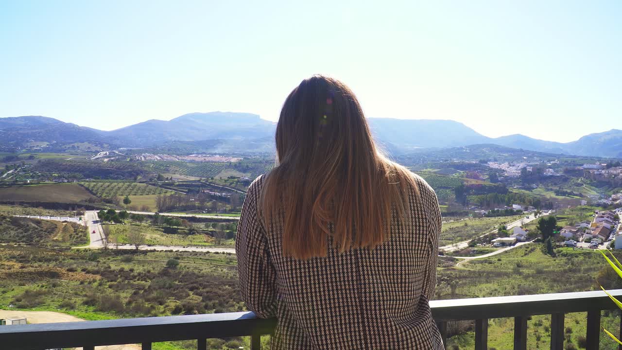 Mädchen mit glatten Haaren, kariertem Mantel und Sonnenbrille, mit dem Rücken zur Kamera, vor einem sonnigen Hintergrund aus Bergen und Feldern