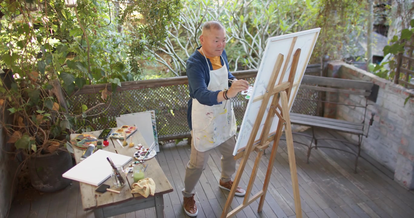 Senior Asian man dipping brush into palette and painting canvas at easel, creating art, copy space