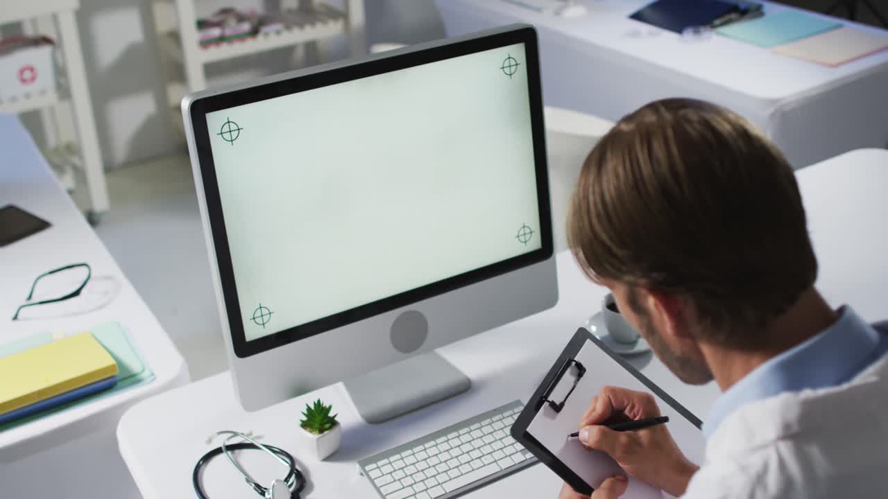 médico caucásico de sexo masculino que realiza una consulta por videollamada utilizando una computadora de escritorio y tomando notas