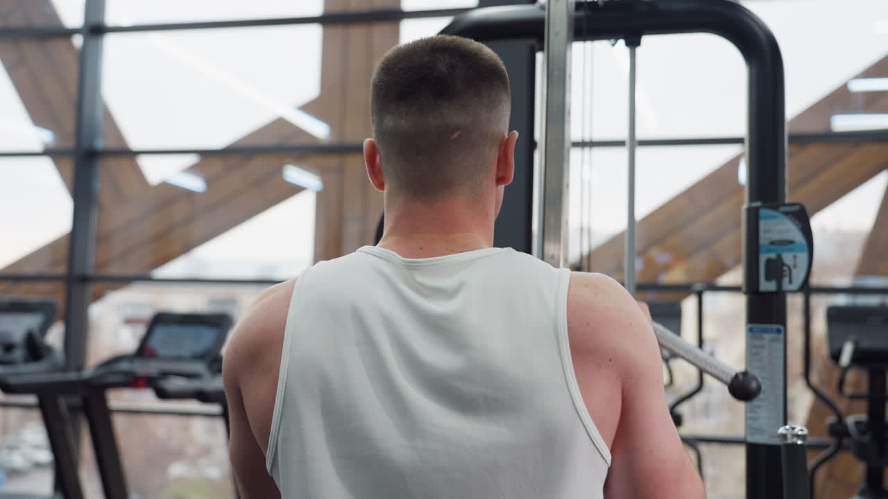 Back view of fitness enthusiast working out facing large window while pulling cable weight machine handle with focused muscle effort and visible strength in spacious modern gym environment