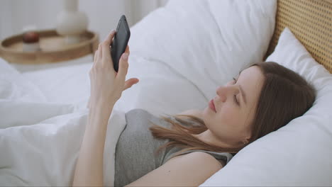 vista de cerca de una mujer escribiendo un mensaje en un teléfono móvil con 2 manos mientras está cómodamente acostada en una cama en una habitación de hotel. mujer usando un teléfono inteligente y acostada en la cama por la mañana después de despertarse